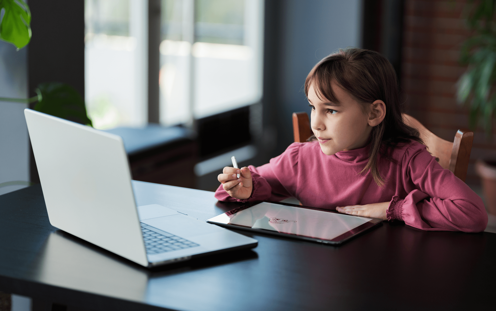 A young girl sits at a table using a stylus on a tablet while looking at a laptop screen, appearing focused.