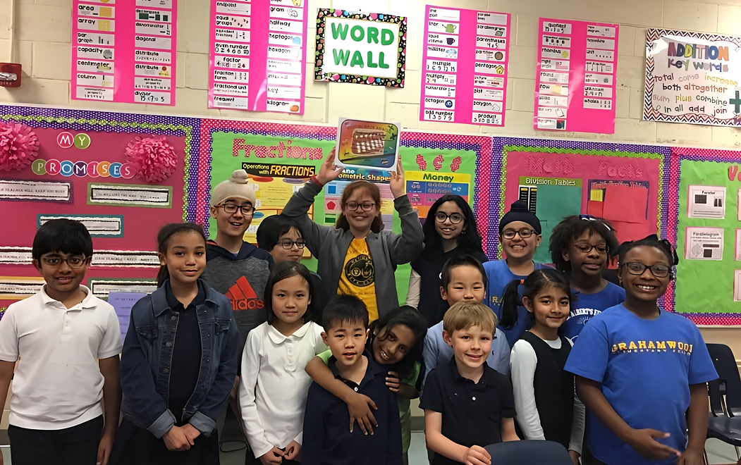 A group of elementary school children and a teacher stand smiling in a colorful classroom with math and reading posters on the walls; one student holds up a small box.