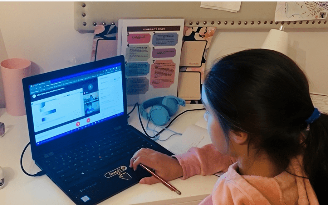 A girl in a pink sweatshirt sits at a desk using a laptop for an online meeting, with headphones, papers, and notes visible around her.