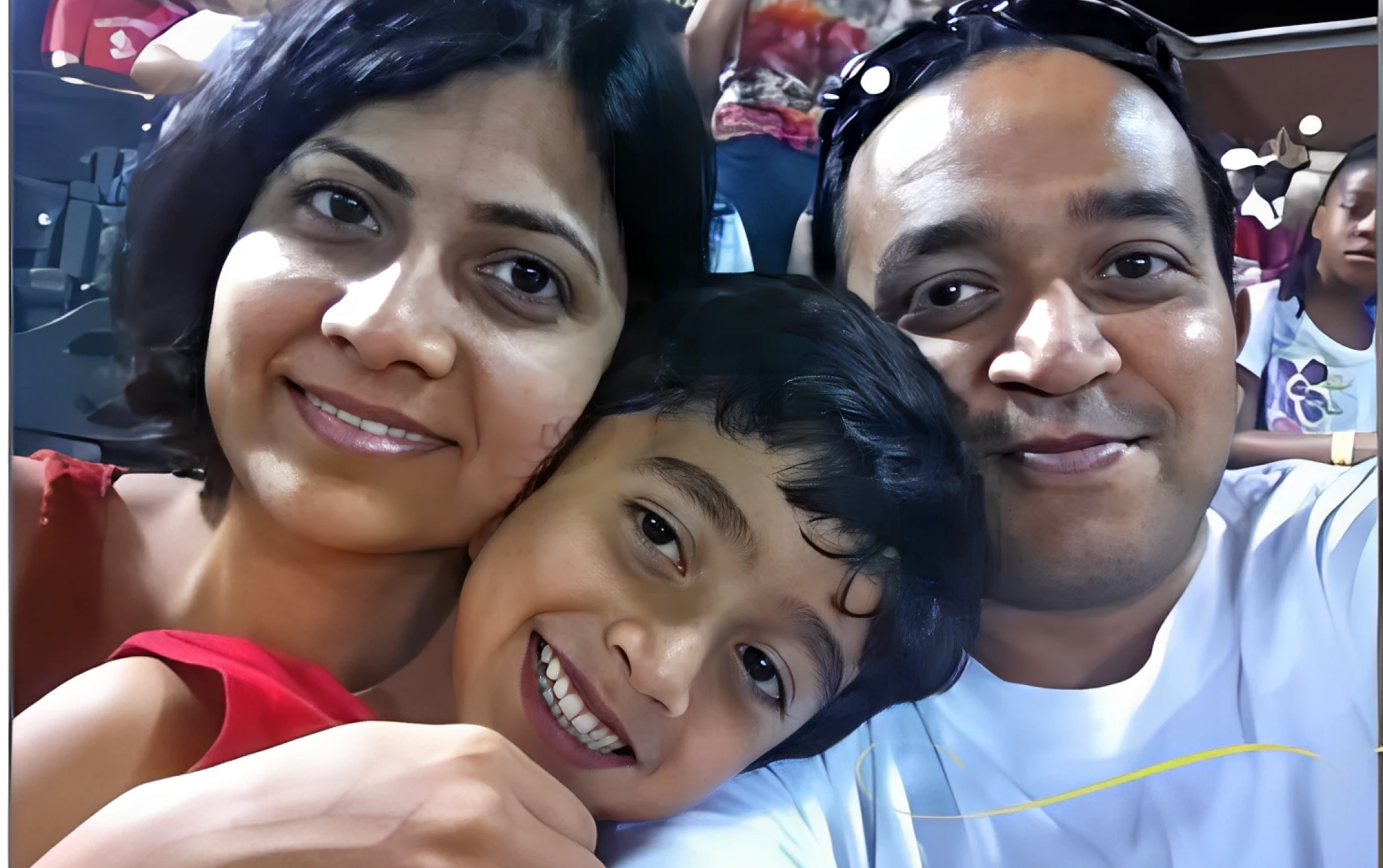 A woman, a child, and a man sit closely together, smiling at the camera in a crowded indoor setting.