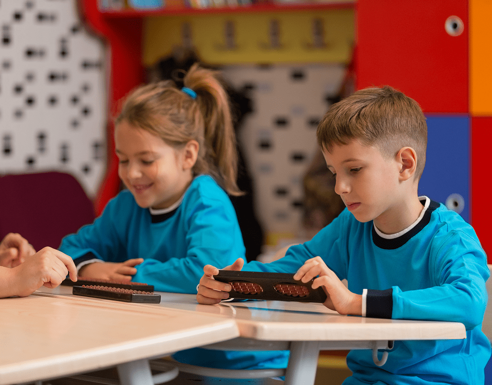 Two children wearing blue shirts sit at a table using abacuses in a classroom setting.