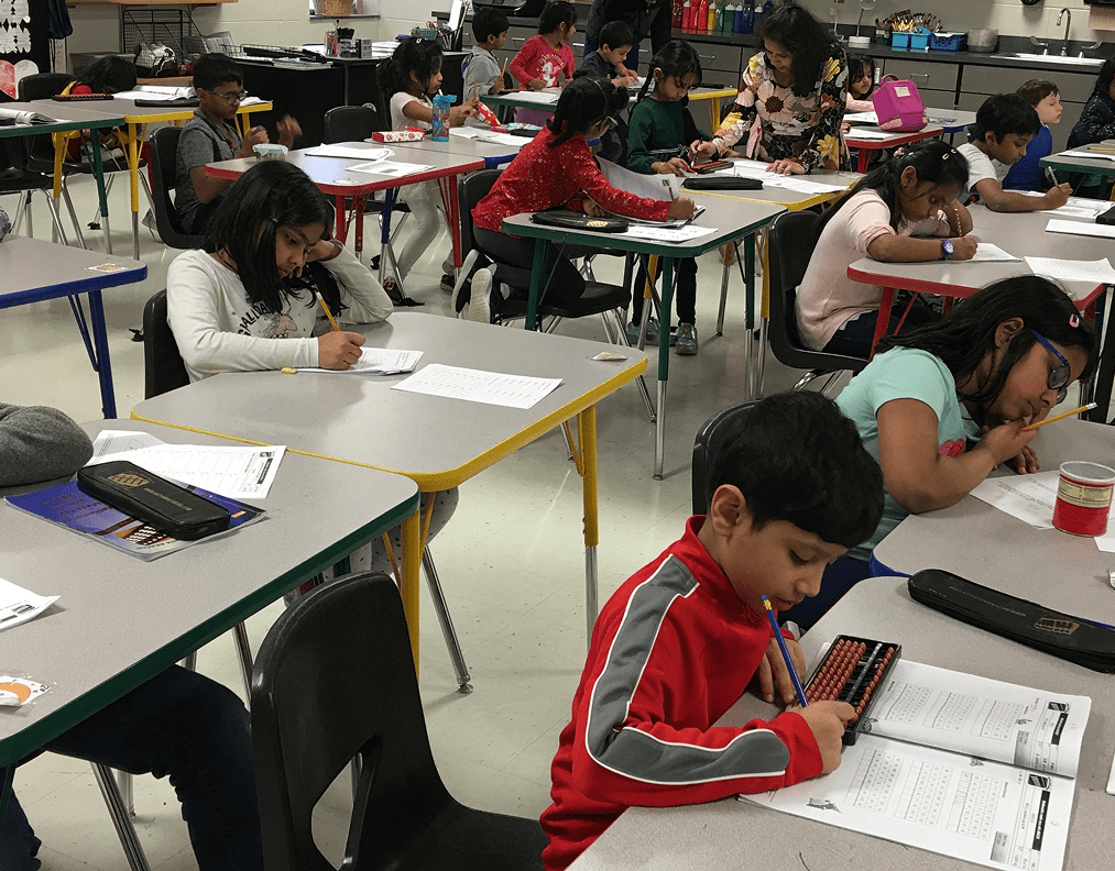Children in a classroom sit at desks working on worksheets, using pencils and abacuses, with a teacher assisting one student.