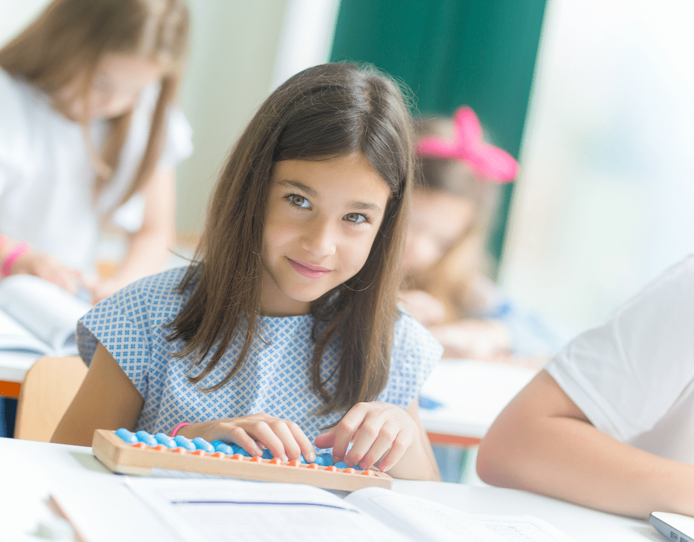 A girl in a classroom uses an abacus, looking at the camera, with other students working in the background.