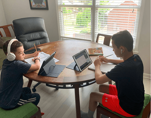 Two boys sit at a round table using tablets and wearing headphones, with notebooks and papers beside them, in a bright room with a large window.
