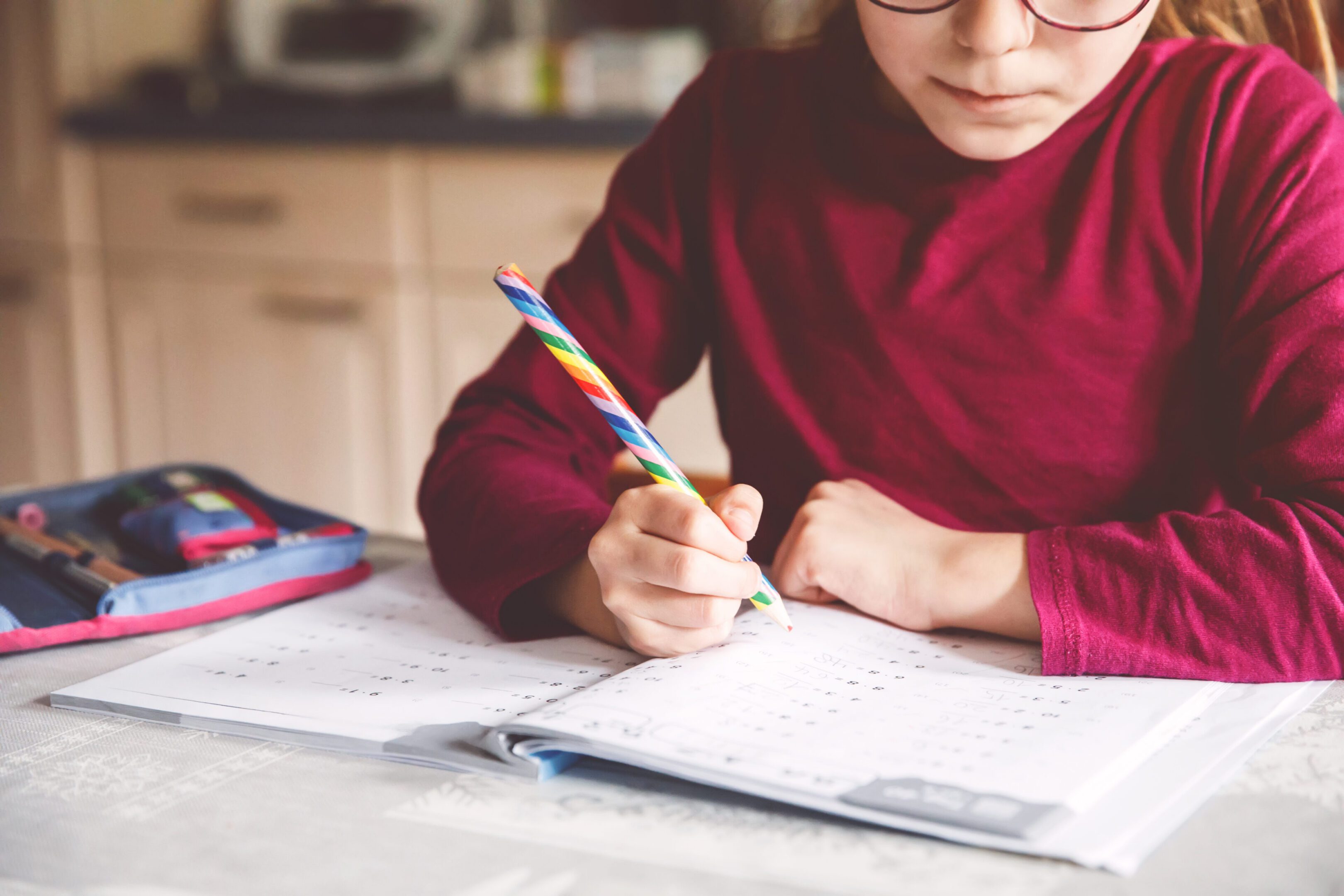 Little school girl learning at home. Elementary class student chid doing maths homework, writing with pencil. Happy pupil with eye glasses.