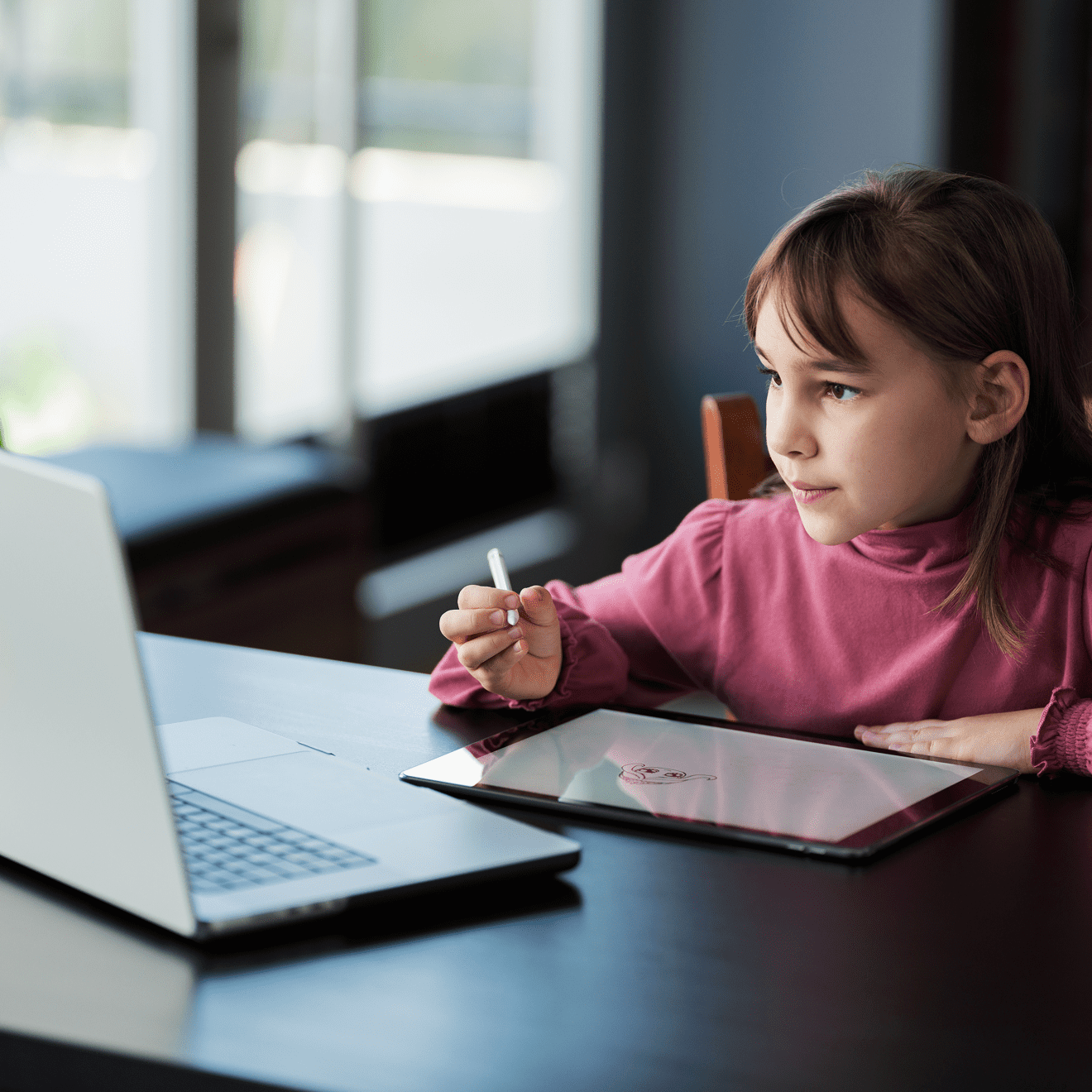 A young girl sits at a table using a stylus on a tablet while looking at a laptop screen, appearing focused.