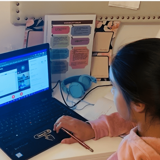 A girl in a pink sweatshirt sits at a desk using a laptop for an online meeting, with headphones, papers, and notes visible around her.